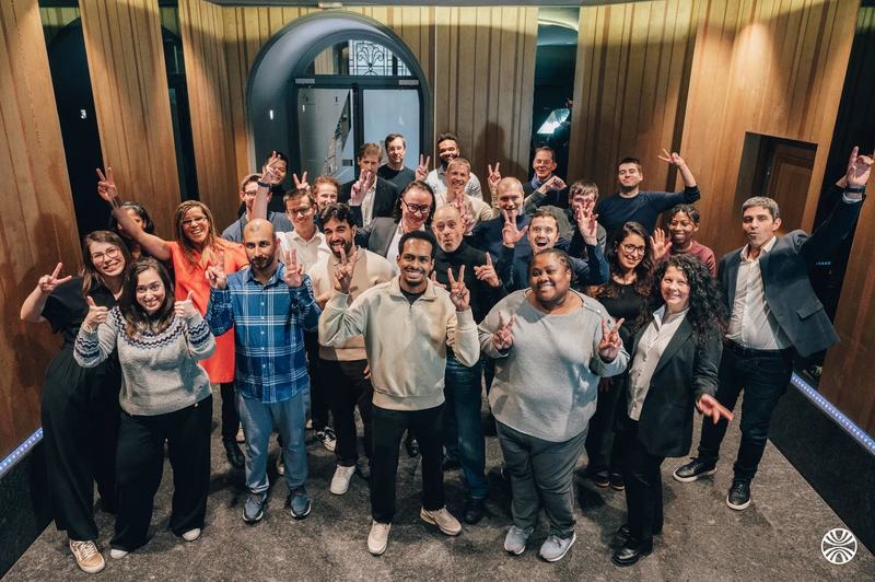 Photo de groupe d’une trentaine de personnes souriantes, nombre d’entre elles faisant le signe de la victoire, dans un hall aux murs boisés.
