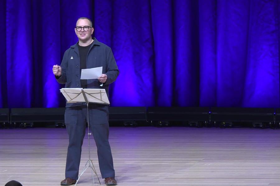 Un homme à lunettes, en veste sombre, se tient sur une scène devant un rideau éclairé en bleu et violet, une feuille à la main, à côté d’un pupitre à partitions.