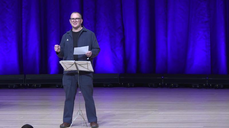 Un homme à lunettes, en veste sombre, se tient sur une scène devant un rideau éclairé en bleu et violet, une feuille à la main, à côté d’un pupitre à partitions.