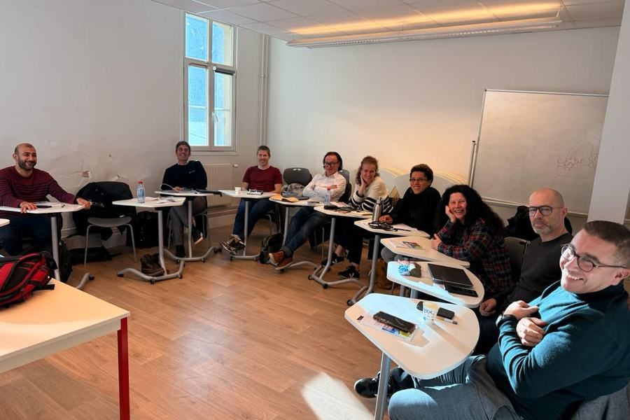 Photo d’une dizaine de personnes souriantes, assises en arc de cercle à des tables écritoires dans une salle de formation, avec un tableau blanc au fond.