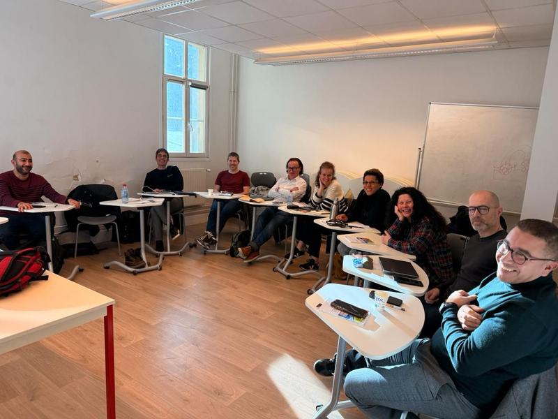 Photo d’une dizaine de personnes souriantes, assises en arc de cercle à des tables écritoires dans une salle de formation, avec un tableau blanc au fond.