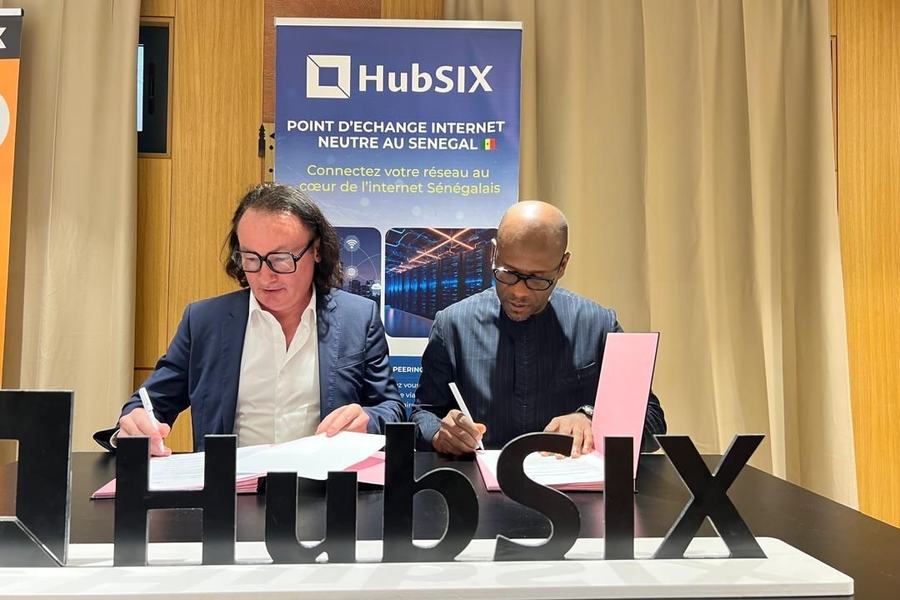 Deux personnes signent des documents à une table, derrière les lettres « HubSIX » et une affiche « HubSIX, point d’échange internet neutre au Sénégal ».