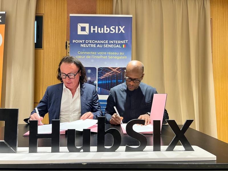 Deux personnes signent des documents à une table, derrière les lettres « HubSIX » et une affiche « HubSIX, point d’échange internet neutre au Sénégal ».