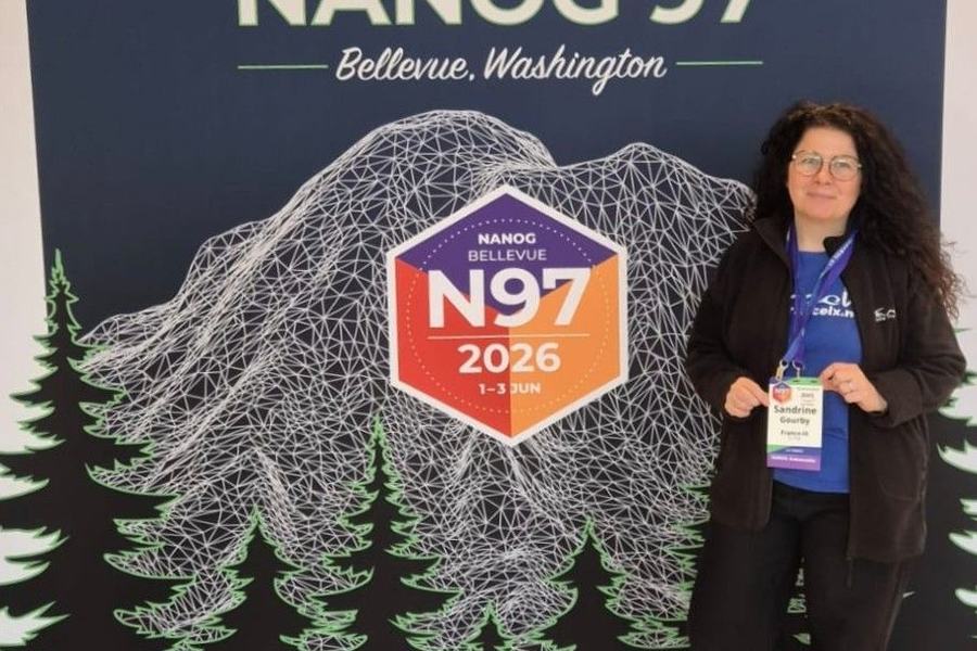 Sandrine Gourby, badge France-IX au cou, pose devant le panneau « NANOG 97, Bellevue, Washington » orné du logo « N97, 2026, 1–3 Jun » et d’une montagne stylisée.