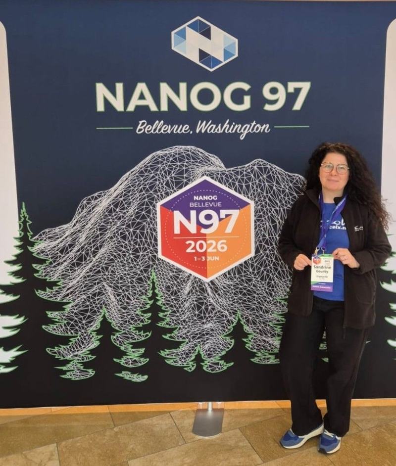 Sandrine Gourby, badge France-IX au cou, pose devant le panneau « NANOG 97, Bellevue, Washington » orné du logo « N97, 2026, 1–3 Jun » et d’une montagne stylisée.