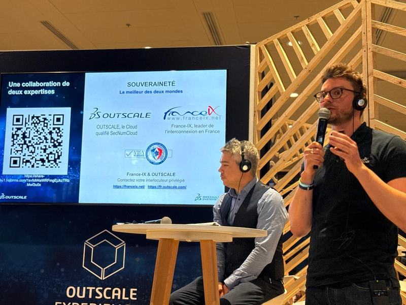 Sur une scène marquée « OUTSCALE », un homme parle au micro à côté d’un homme assis, devant un écran qui affiche « Une collaboration de deux expertises », « Souveraineté, le meilleur des deux mondes » et les logos d’OUTSCALE et de France-IX.