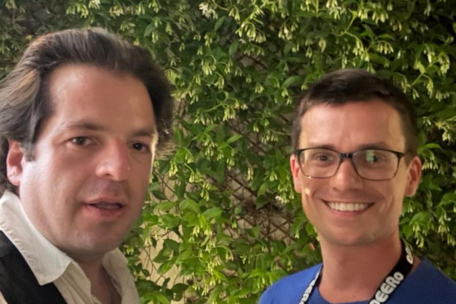 Selfie de deux hommes devant un mur de plantes grimpantes en fleurs ; l’un porte un t-shirt bleu imprimé du logo France-IX et de l’adresse www.franceix.net, avec un tour de cou marqué « aPEERo ».