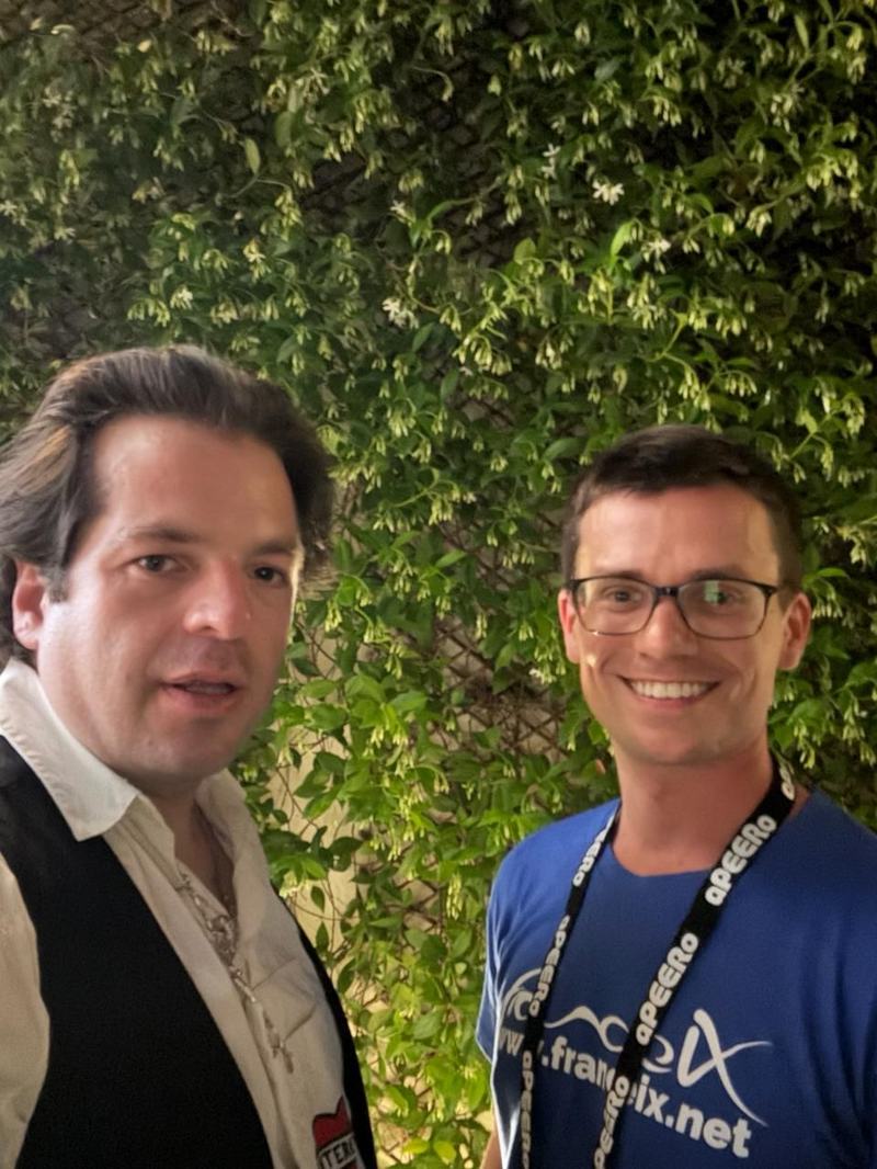 Selfie de deux hommes devant un mur de plantes grimpantes en fleurs ; l’un porte un t-shirt bleu imprimé du logo France-IX et de l’adresse www.franceix.net, avec un tour de cou marqué « aPEERo ».