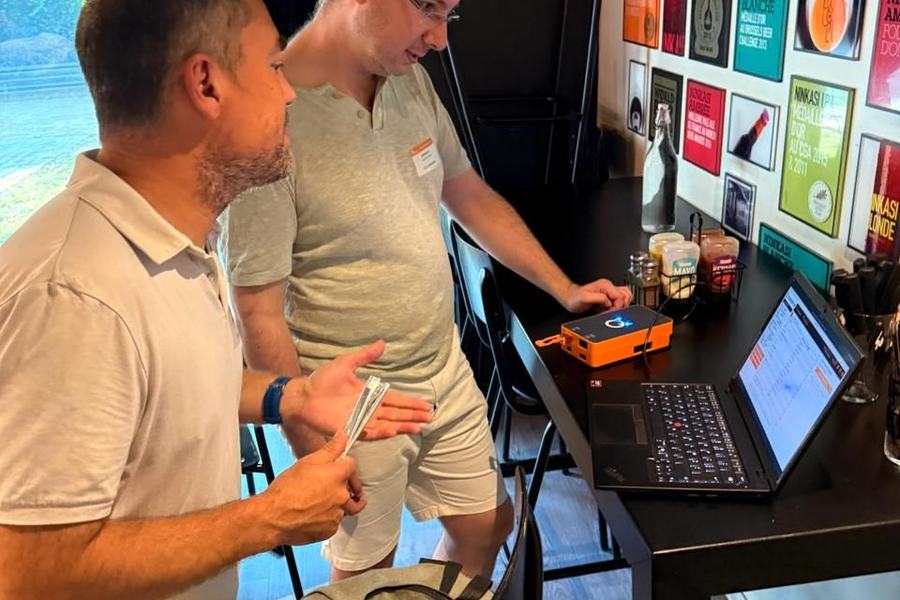 Deux hommes debout devant une table haute regardent l’écran d’un ordinateur portable, à côté d’un petit boîtier orange ; au mur, des affiches encadrées de bières Ninkasi et une affiche « World Beer Awards ».