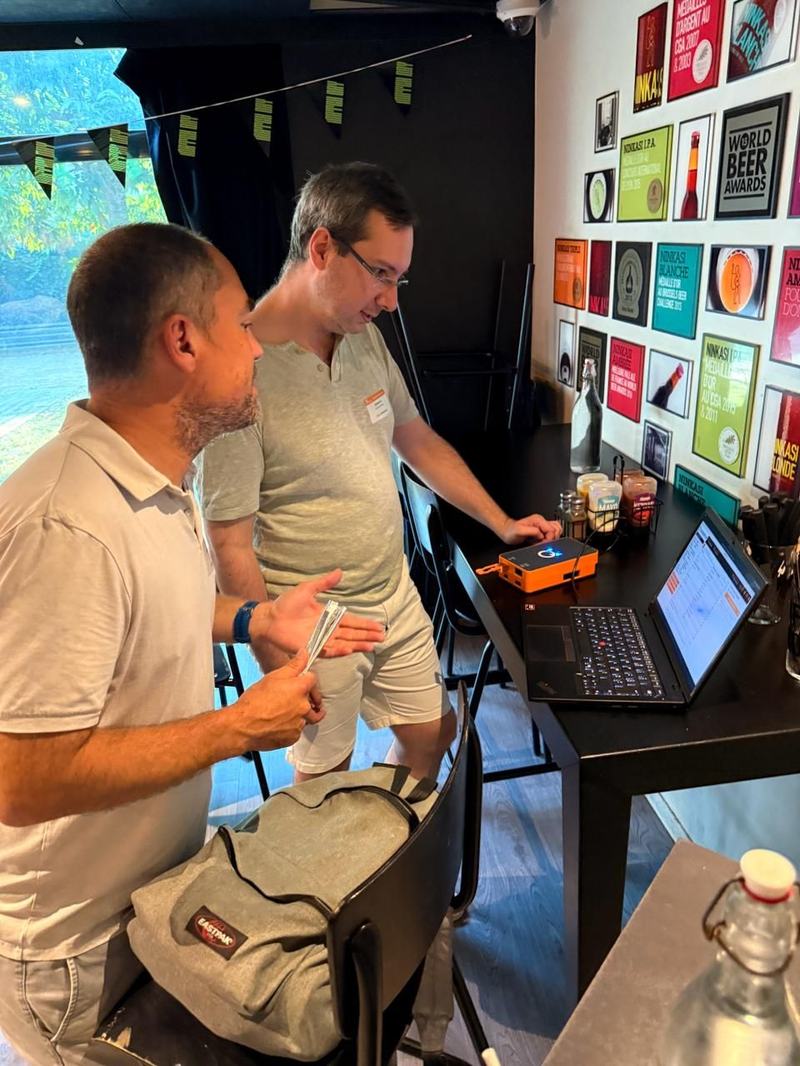 Deux hommes debout devant une table haute regardent l’écran d’un ordinateur portable, à côté d’un petit boîtier orange ; au mur, des affiches encadrées de bières Ninkasi et une affiche « World Beer Awards ».