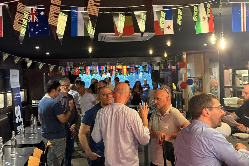 Des participants discutent debout dans une salle décorée de guirlandes de drapeaux, à côté d’un kakémono France-IX.