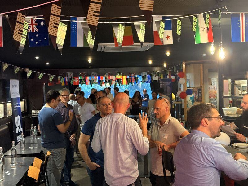 Des participants discutent debout dans une salle décorée de guirlandes de drapeaux, à côté d’un kakémono France-IX.
