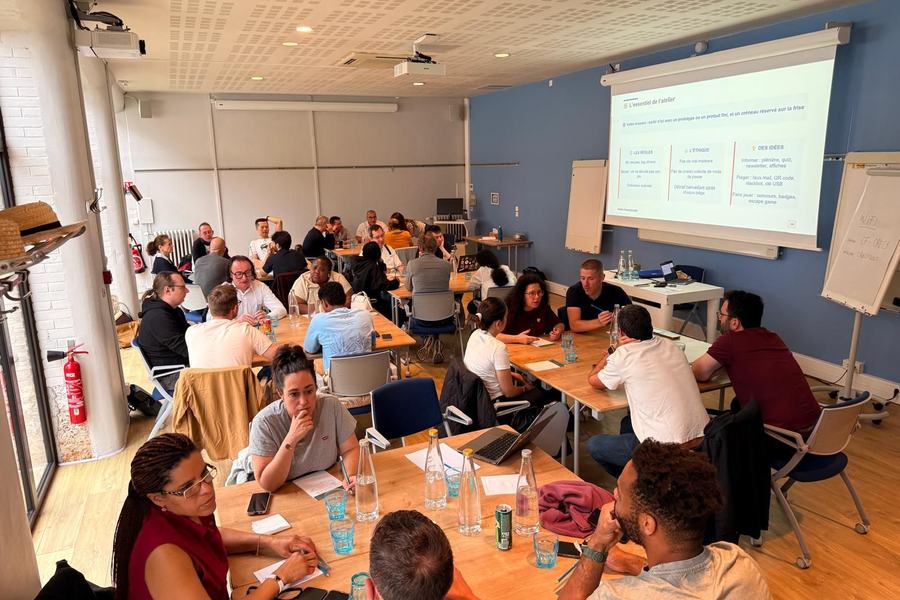 Atelier en salle : de nombreuses personnes travaillent en groupes autour de tables, devant une diapositive « L’essentiel de l’atelier » qui détaille les règles, l’éthique (« Pas de vrai malware ») et des idées de pièges.