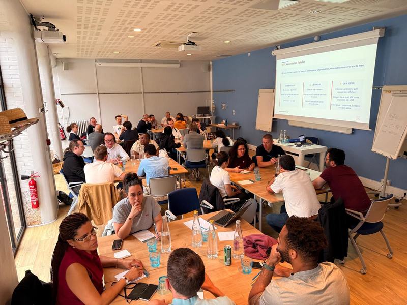 Atelier en salle : de nombreuses personnes travaillent en groupes autour de tables, devant une diapositive « L’essentiel de l’atelier » qui détaille les règles, l’éthique (« Pas de vrai malware ») et des idées de pièges.