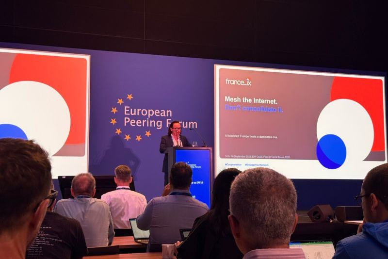 Salle de conférence de l’European Peering Forum : un intervenant au pupitre ; à l’écran, la diapositive France-IX « Mesh the Internet. Don’t consolidate it. », au nom de Franck Simon, CEO.