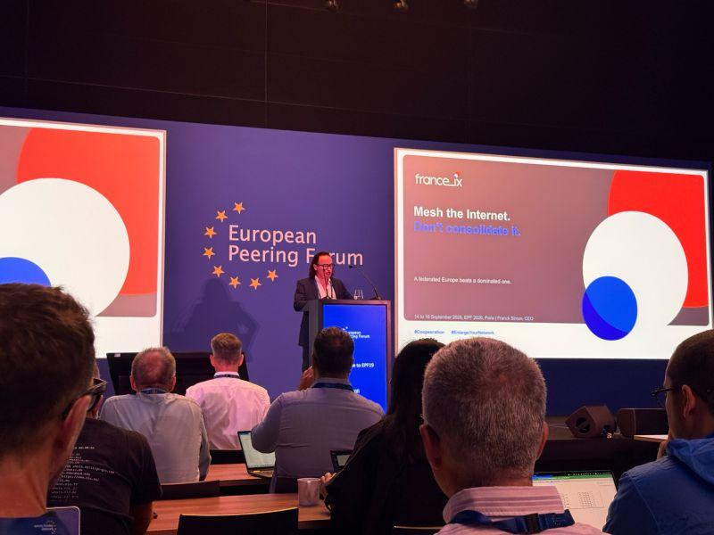 Salle de conférence de l’European Peering Forum : un intervenant au pupitre ; à l’écran, la diapositive France-IX « Mesh the Internet. Don’t consolidate it. », au nom de Franck Simon, CEO.