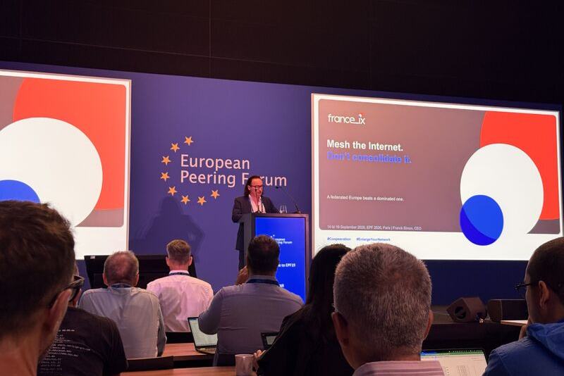 Salle de conférence de l’European Peering Forum : un intervenant au pupitre ; à l’écran, une diapositive France-IX « Mesh the Internet. Don’t consolidate it. ».