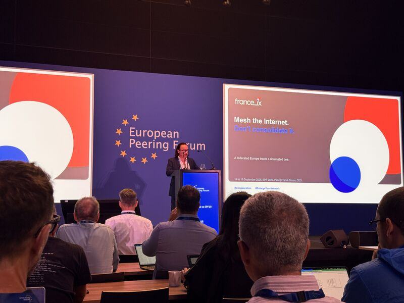 Salle de conférence de l’European Peering Forum : un intervenant au pupitre ; à l’écran, une diapositive France-IX « Mesh the Internet. Don’t consolidate it. ».