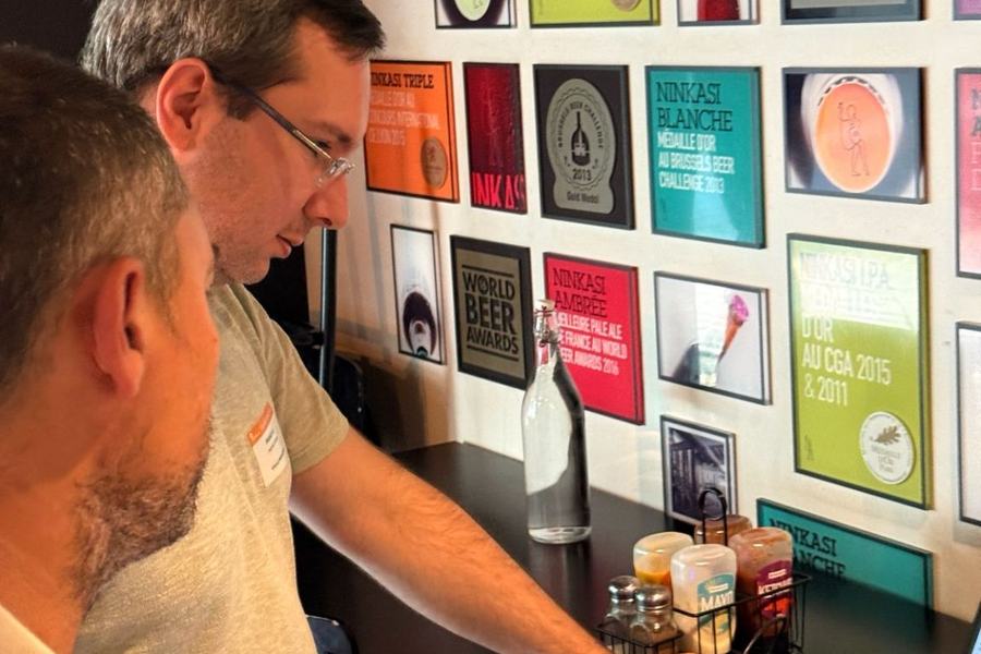 Deux hommes devant un mur d’affiches encadrées de bières Ninkasi et des « World Beer Awards » ; l’un appuie sur le bouton d’un boîtier orange relié à un ordinateur portable.