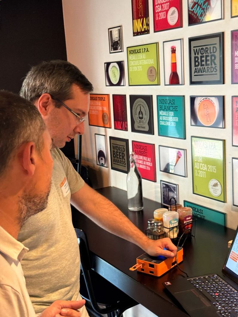 Deux hommes devant un mur d’affiches encadrées de bières Ninkasi et des « World Beer Awards » ; l’un appuie sur le bouton d’un boîtier orange relié à un ordinateur portable.